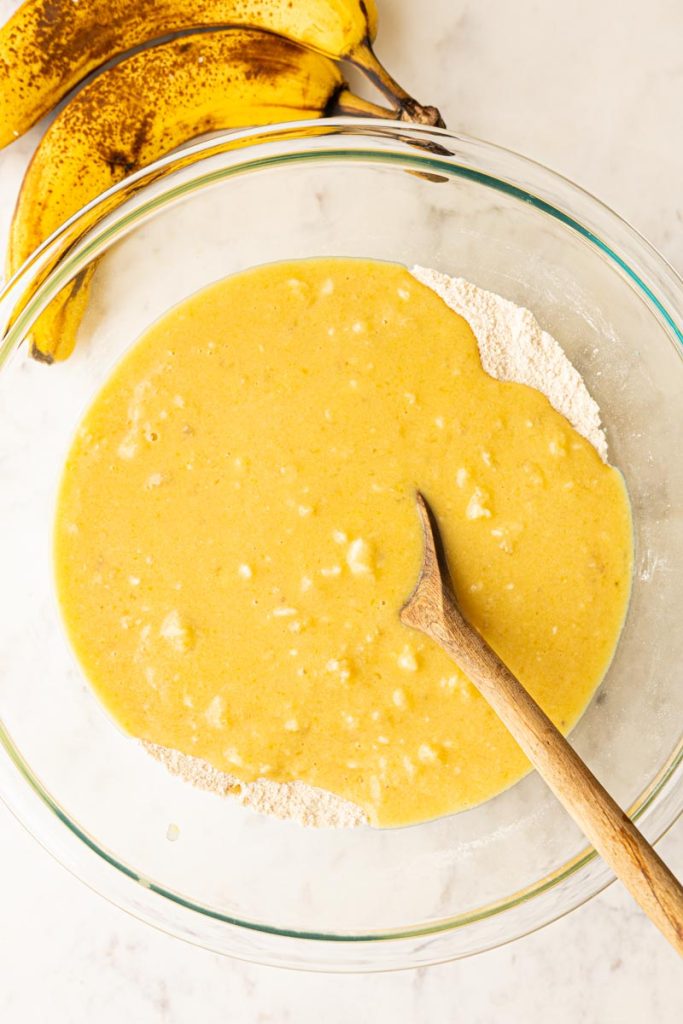 Banana bread batter in a glass mixing bowl with a wooden spoon.