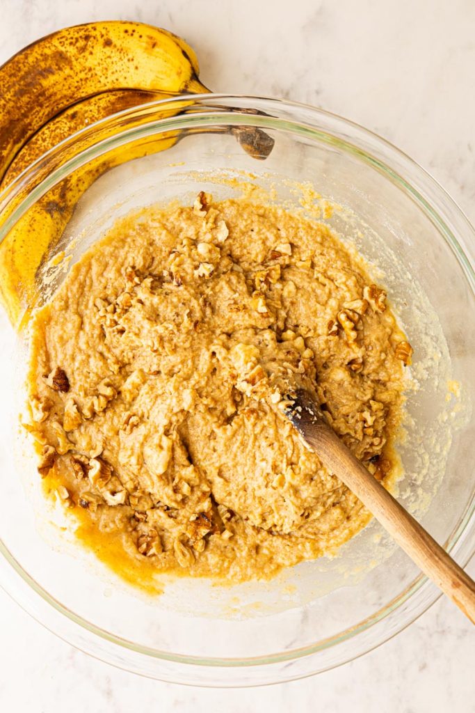 Banana bread batter with chopped walnuts in a glass mixing bowl with a wooden spoon.