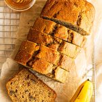 A sliced loaf of healthy banana bread on a piece of parchment paper and cooling rack. A jar of honey and two ripe bananas are next to the bread.