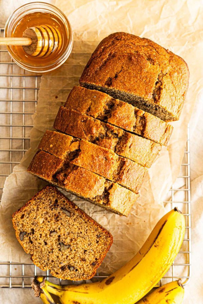 A sliced loaf of healthy banana bread on a piece of parchment paper and cooling rack. A jar of honey and two ripe bananas are next to the bread.