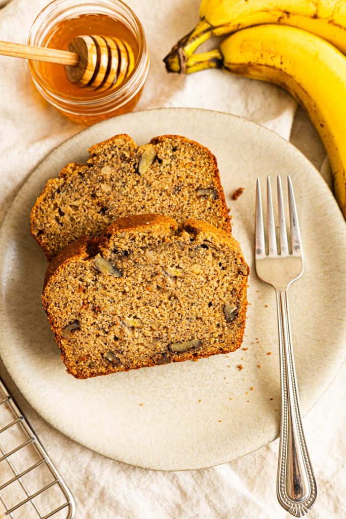 Two slices of healthy banana bread on a plate with a fork. A jar of honey and 2 ripe bananas next to the plate.