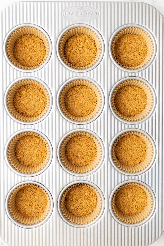 Baked graham cracker crust for mini cheesecakes, inside muffin liners in a baking pan.