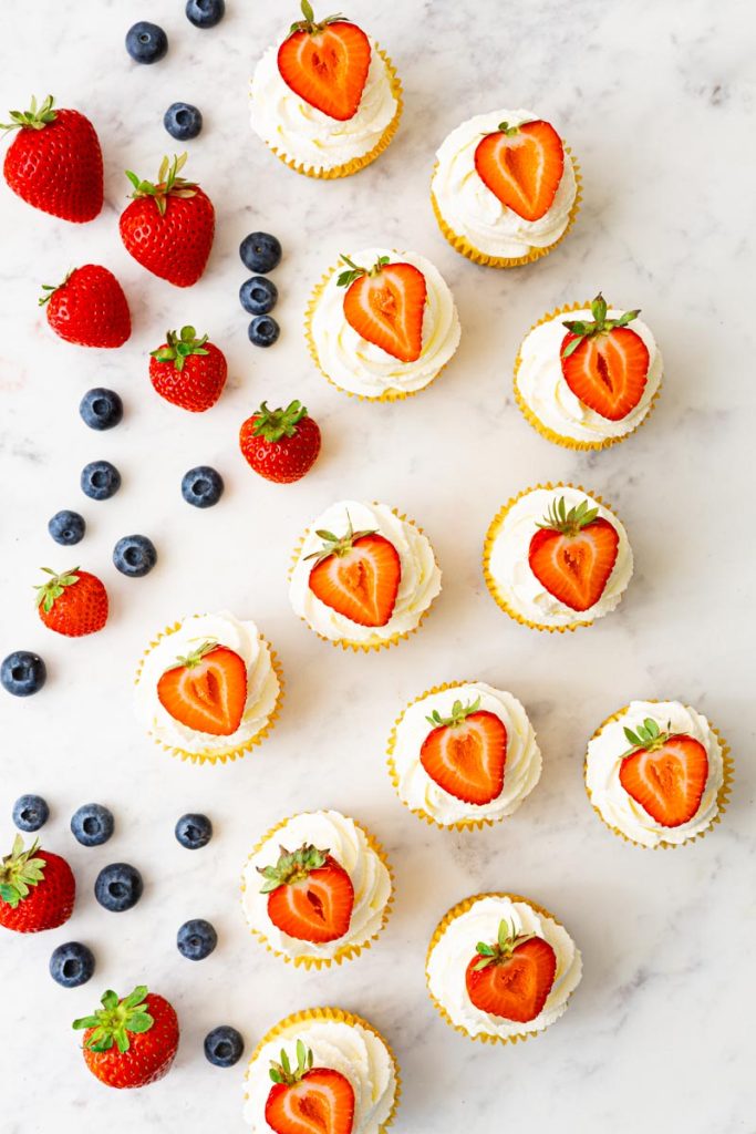 Mini cheesecakes decorated with a whipped cream topping and half a strawberry. Strawberries and blueberries are around the cheesecakes.