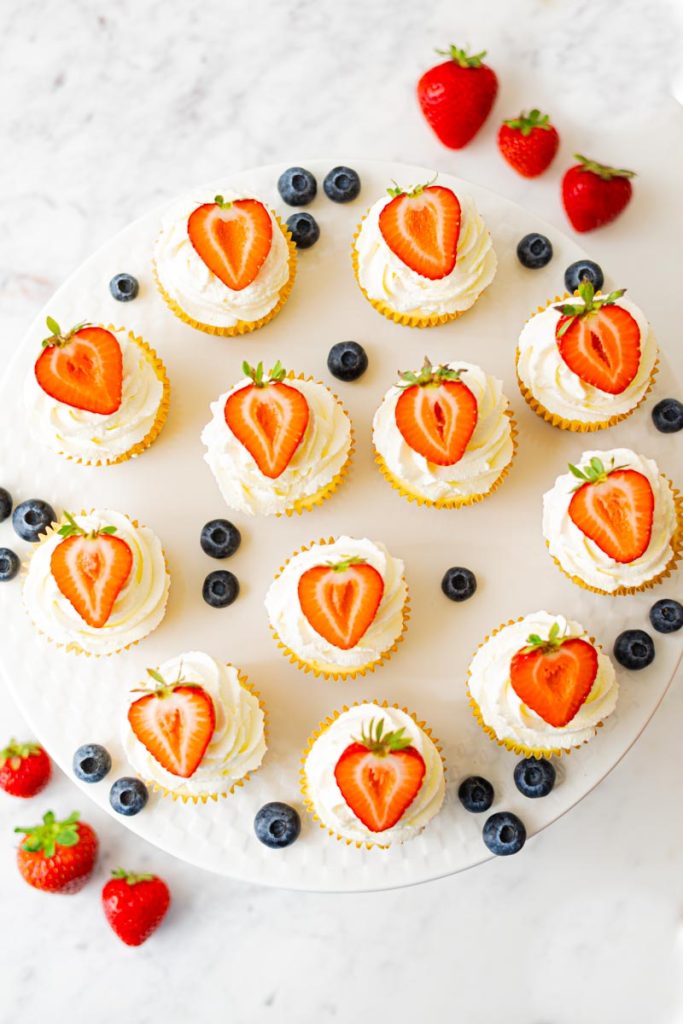 Cake stand with mini cheesecakes, decorated with a whipped cream topping and half a strawberry. Strawberries and blueberries are around the cheesecakes.