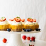 Cake stand with mini cheesecakes, decorated with a whipped cream topping and half a strawberry. Strawberries and blueberries are around the cheesecakes.