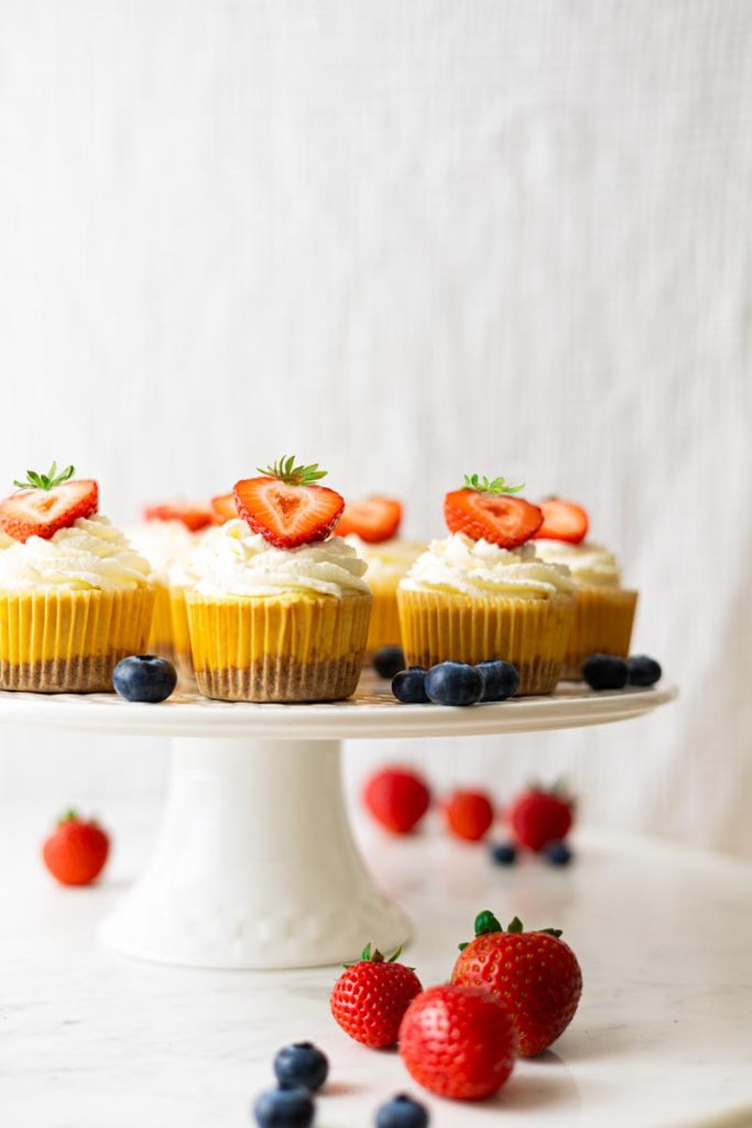 Cake stand with mini cheesecakes, decorated with a whipped cream topping and half a strawberry. Strawberries and blueberries are around the cheesecakes.