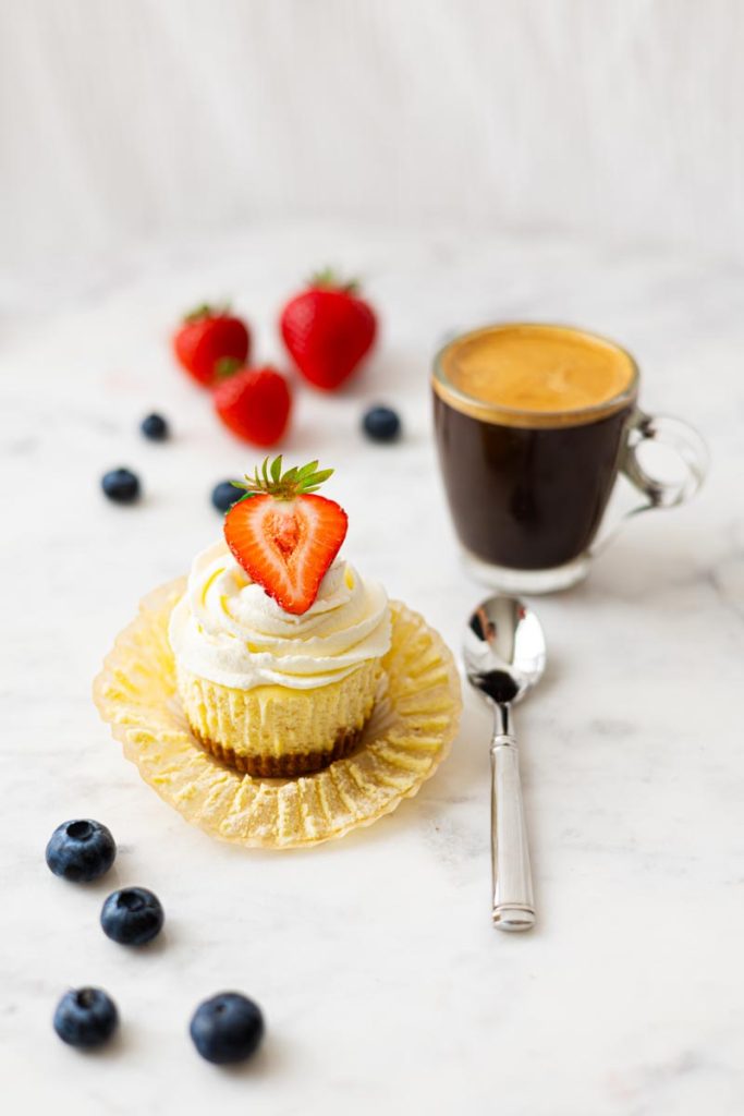 Mini cheesecake with a strawberry on top, with a spoon and a cup of coffee.