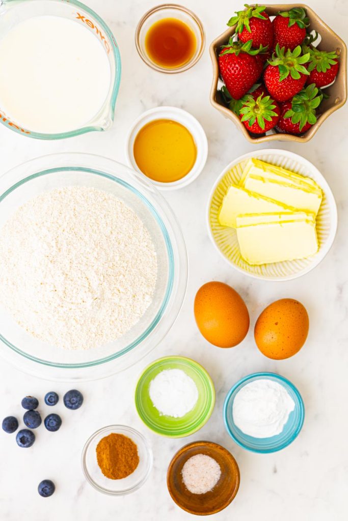 Ingredients to make fluffy whole wheat waffles: whole wheat flour, buttermilk, eggs, butter, honey, vanilla, baking soda, baking powder, cinnamon, and salt. A colander of strawberries and a couple scattered blueberries.