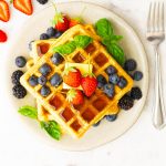 A plate with fluffy whole wheat waffles, topped with butter, maple syrup, strawberries, blueberries, blackberries and basil as garnish.
