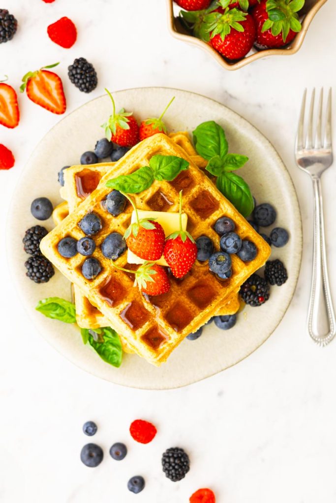 A plate with fluffy whole wheat waffles, topped with butter, maple syrup, strawberries, blueberries, blackberries and basil as garnish.