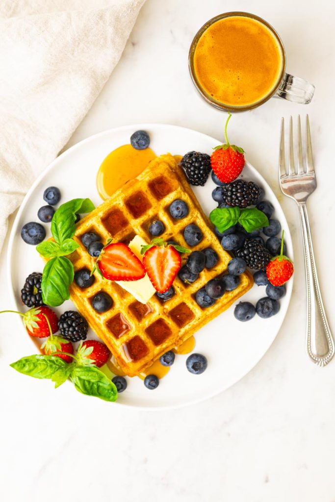 A plate with fluffy whole wheat waffles, topped with butter, maple syrup, strawberries, blueberries, blackberries and basil as garnish. A cup of coffee.