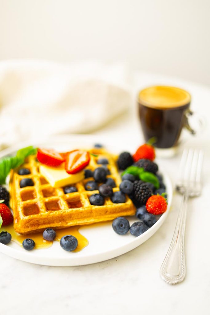 A plate with fluffy whole wheat waffles, topped with butter, maple syrup, strawberries, blueberries, blackberries and basil as garnish. A cup of coffee.