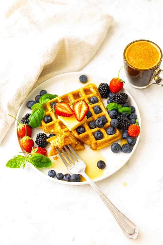 A plate with fluffy whole wheat waffles, topped with butter, maple syrup, strawberries, blueberries, blackberries and basil as garnish. A cup of coffee.