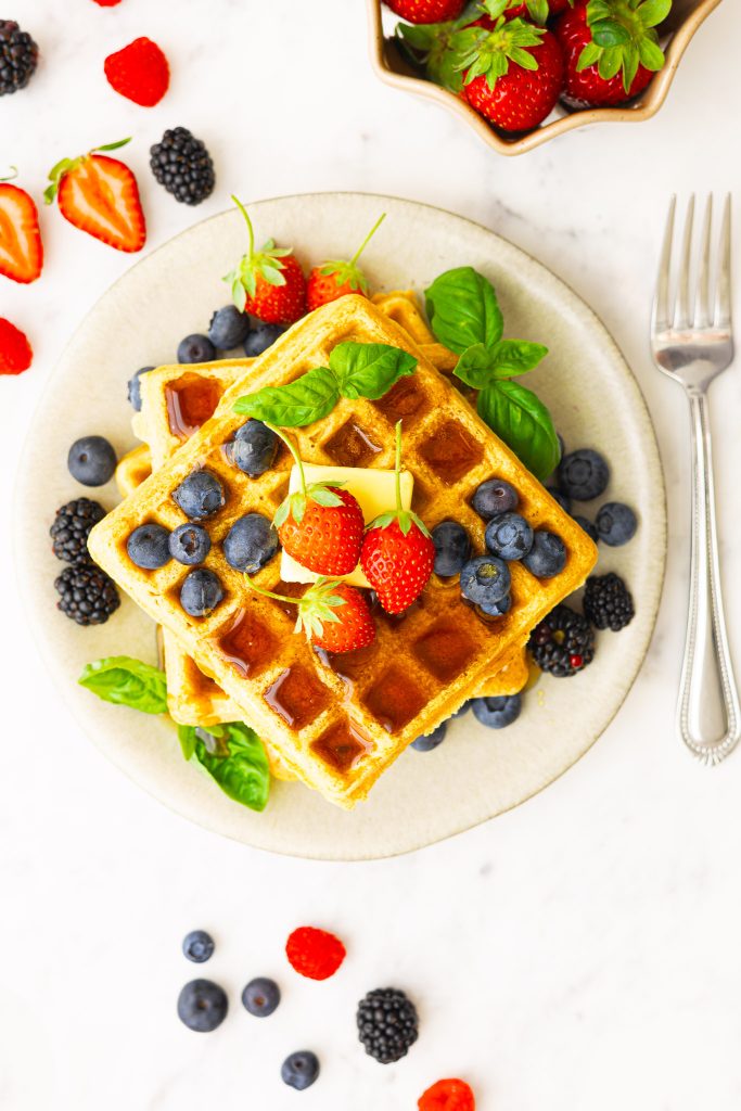 A plate with fluffy whole wheat waffles, topped with butter, maple syrup, strawberries, blueberries, blackberries and basil as garnish.